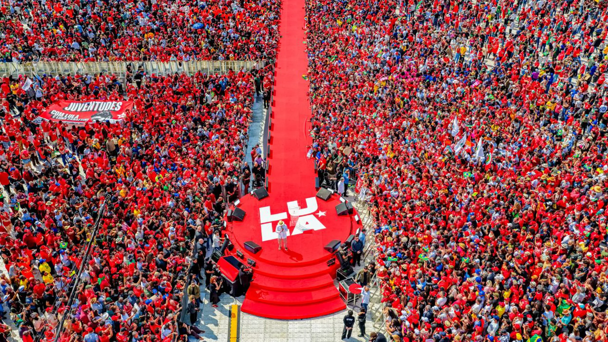 Supporters in red fill a crowd around a stage with the Lula logo