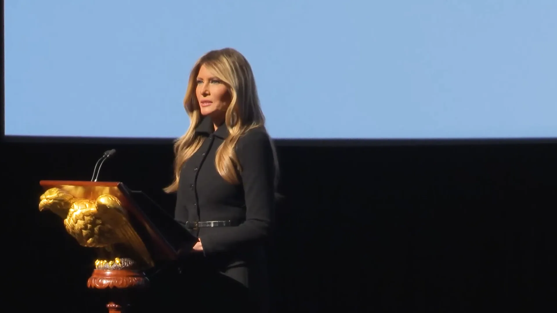 Melania Trump speaking at a podium on a dark stage